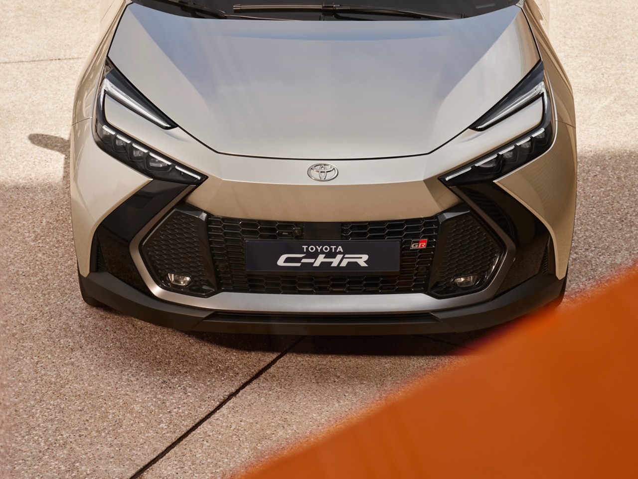 The front view of a New Toyota C-HR in silver with a black grille. It's parked on stone paving with a flash of orange in the foreground.