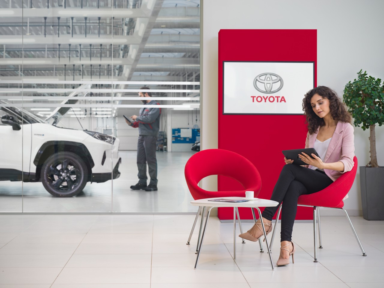 Une femme est assise dans la salle d’attente d’un centre de service Toyota. Elle regarde sa tablette. Derrière elle, le logo Toyota s’affiche sur un écran de télévision.