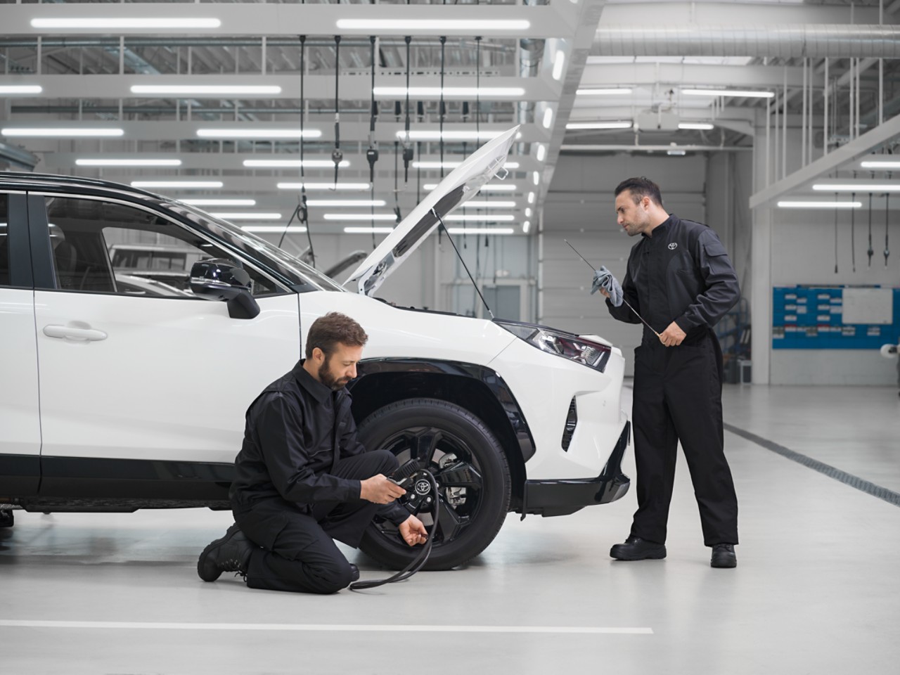 Deux techniciens s’occupent de l’entretien d’un Toyota RAV4 blanc dont le capot est levé. L’un est agenouillé et vérifie la pression des pneus, l’autre contrôle le niveau d’huile.