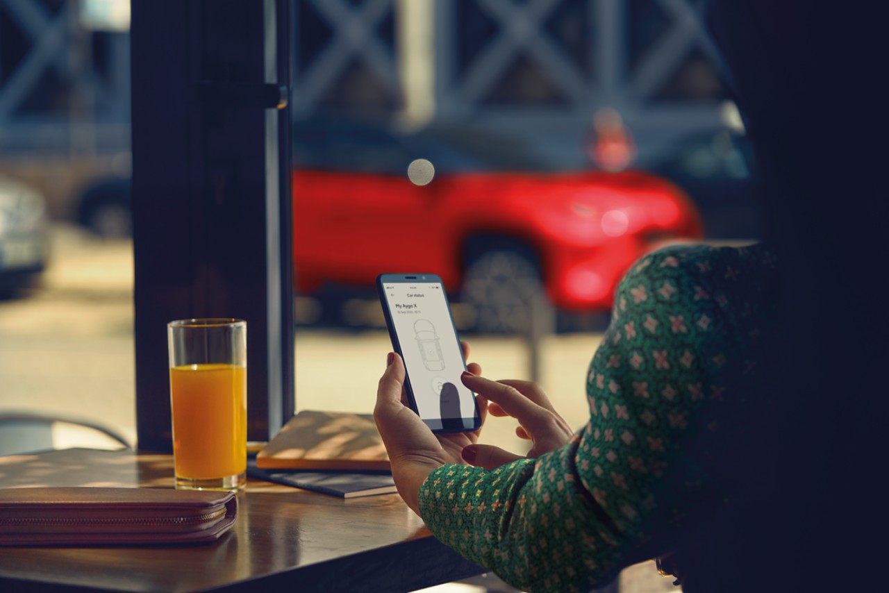 Une femme assise dans un café utilise l’appli MyT de Toyota sur son téléphone. Son Toyota Yaris Cross est parqué en arrière-plan.
