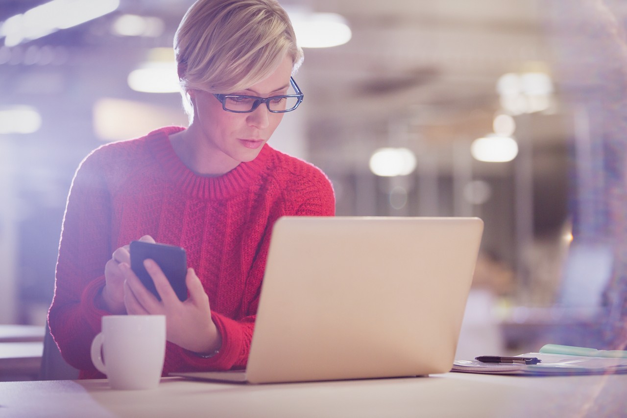 Une femme portant des lunettes et un pull rouge regarde son ordinateur portable, son smartphone à la main. Une tasse de café est posée sur la table.